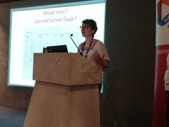 Dr Alexandra Mitchell at the CAHG 2019 Conference.. A person stands behind a lectern gesticulating, a powerpoint is behind them.