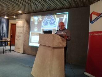 Harvey Kaplan at the CAHG 2019 Conference.. A person stands behind a lectern gesticulating, a powerpoint is behind them.