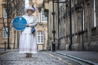 Sarah Cobham, in period costume, holding a blue plaque, near Wakefield town hall, with the blue plaque dedicated to Mary Francis Heaton; West Riding Pauper Lunatic Asylum, in 1818