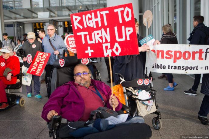 A man using a powerchair holds up a red protest sign that reads 